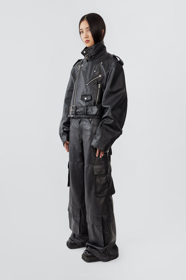 A female model wears the Dormi Oversized Leather Jacket in black by Deadwood. This bold, cropped biker jacket is crafted from heavy upcycled cowskin and features a boxy, oversized fit that sits just above the waist. Classic biker details include an asymmetrical front zipper, a chunky belt, a coin pocket, shoulder epaulets, and zippered cuffs and pockets. The lining is made from 100% recycled PET bottles, combining edgy style with sustainability.
