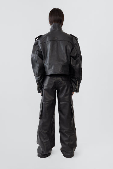 A female model wears the Dormi Oversized Leather Jacket in black by Deadwood. This bold, cropped biker jacket is crafted from heavy upcycled cowskin and features a boxy, oversized fit that sits just above the waist. Classic biker details include an asymmetrical front zipper, a chunky belt, a coin pocket, shoulder epaulets, and zippered cuffs and pockets. The lining is made from 100% recycled PET bottles, combining edgy style with sustainability.