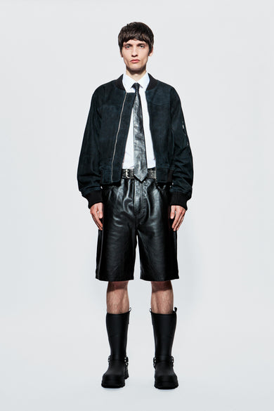A male model wearing a dark panel leather bomber jacket with a round neckline, zipper closure, waist pockets, and ribbed collar, cuffs, and hem. The model is also wearing a white shirt, black boots, and the jacket appears to be black in color.