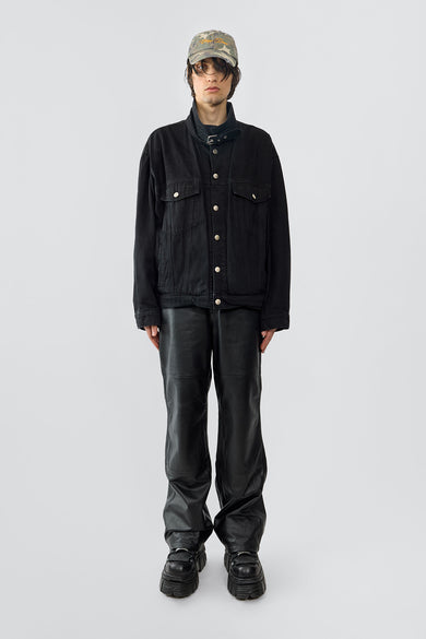 The Oracle Denim Jacket is an oversized black cotton denim jacket with steel jean buttons, flap chest pockets, and waist pockets. It features a strap and buckle at the collar for a uniform-inspired look, with dropped shoulders for a relaxed fit. Crafted with sustainable fashion in mind.