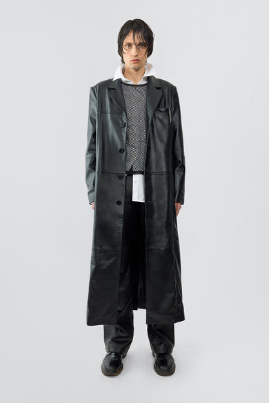 A model wears the Vargen Coat in black, an ultra-long, oversized coat crafted from soft upcycled lambskin. It features a front button closure, welt side pockets, an inside pocket, and a lining made from 100% recycled plastic bottles.
