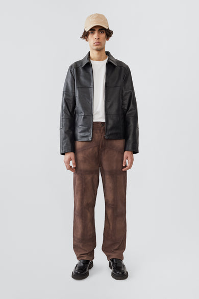 A male model wearing the Bruno Patch Jacket by Deadwood, a straight-cut leather jacket crafted from heavy upcycled cowskin. The design features subtle yet distinct patchwork details, a casual fit, and a waist-length silhouette. Functional elements include a front zipper closure, zippered wrist openings, and a zippered pocket. Lined with fabric made from 100% recycled plastic bottles, this jacket merges timeless craftsmanship with sustainable materials.