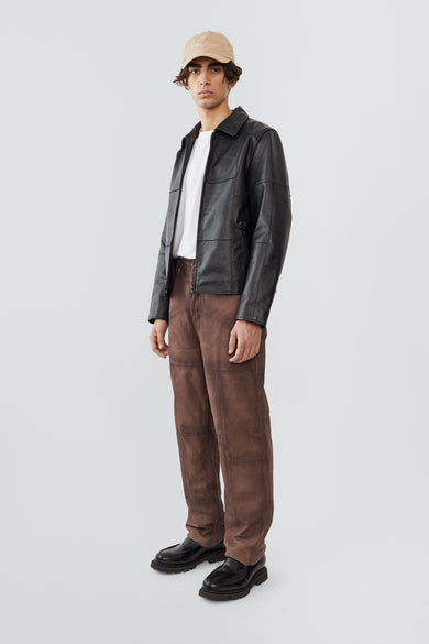A male model wearing the Bruno Patch Jacket by Deadwood, a straight-cut leather jacket crafted from heavy upcycled cowskin. The design features subtle yet distinct patchwork details, a casual fit, and a waist-length silhouette. Functional elements include a front zipper closure, zippered wrist openings, and a zippered pocket. Lined with fabric made from 100% recycled plastic bottles, this jacket merges timeless craftsmanship with sustainable materials.