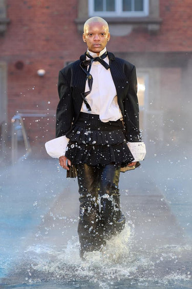 Look 7 from Deadwood FW25 Runway Show at Copenhagen Fashion Week. Model walking on a runway in the water at Villa CPH wearing black pinstriped blazer and skirt, white shirt, black pants in upcycled leather and Tretorn rubber boots.