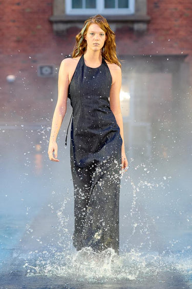 Look 11 from Deadwood FW25 Runway Show at Copenhagen Fashion Week. Model walking on a runway in the water at Villa CPH wearing a black dress in upcycled leather and Tretorn rubber boots.