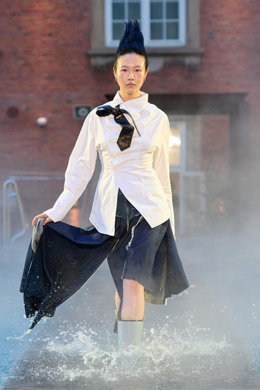 Look 12 from Deadwood FW25 Runway Show at Copenhagen Fashion Week. Model walking on a runway in the water at Villa CPH wearing white shirt made out of recycled cotton and navy skirt in upcycled leather and Tretorn rubber boots.