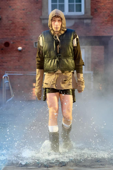 Look 13 from Deadwood FW25 Runway Show at Copenhagen Fashion Week. Model walking on a runway in the water at Villa CPH wearing black shorts and olive vest in upcycled leather and Tretorn rubber boots.