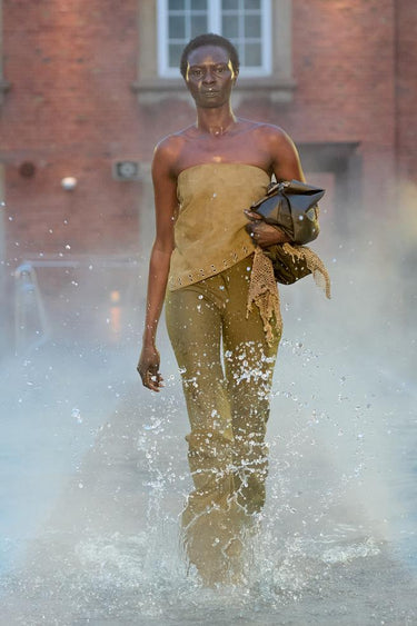 Look 15 from Deadwood FW25 Runway Show at Copenhagen Fashion Week. Model walking on a runway in the water at Villa CPH wearing olive pants and olive top in upcycled leather and Tretorn rubber boots.