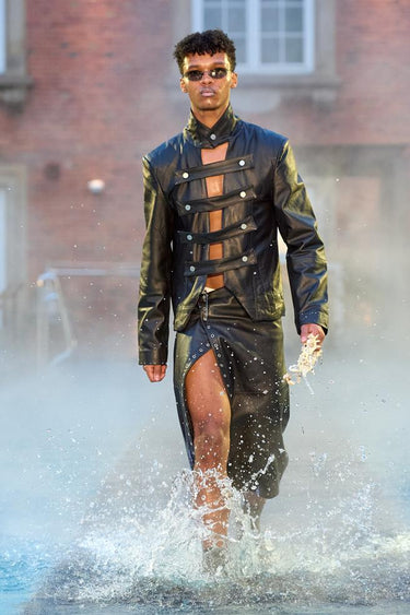 Look 16 from Deadwood FW25 Runway Show at Copenhagen Fashion Week. Model walking on a runway in the water at Villa CPH wearing black skirt and black jacket in upcycled leather and Tretorn rubber boots.