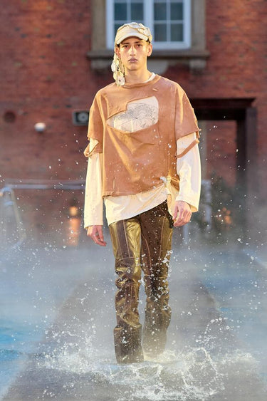 Look 17 from Deadwood FW25 Runway Show at Copenhagen Fashion Week. Model walking on a runway in the water at Villa CPH wearing brown pants in upcycled leather white shirt made of recyled cotton and Tretorn rubber boots.