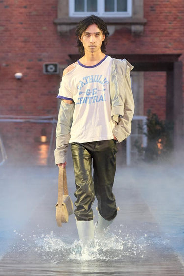 Look 18 from Deadwood FW25 Runway Show at Copenhagen Fashion Week. Model walking on a runway in the water at Villa CPH wearing black pants and grey jacket in upcycled leather and Tretorn rubber boots.