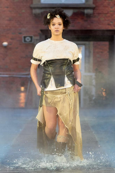 Look 19 from Deadwood FW25 Runway Show at Copenhagen Fashion Week. Model walking on a runway in the water at Villa CPH wearing beige skirt and black top in upcycled leather and Tretorn rubber boots.