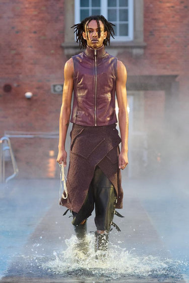 Look 2 from Deadwood FW25 Runway Show at Copenhagen Fashion Week. Model walking on a runway in the water at Villa CPH wearing black pants and burgundy vest in upcycled leather and Tretorn rubber boots.