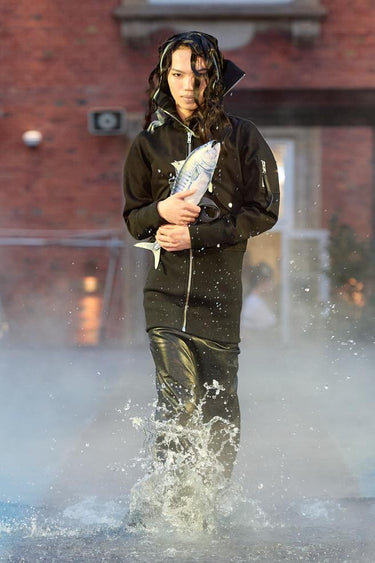 Look 21 from Deadwood FW25 Runway Show at Copenhagen Fashion Week. Model walking on a runway in the water at Villa CPH wearing black pants and black jacket in upcycled leather, a fishshaped bag and Tretorn rubber boots.