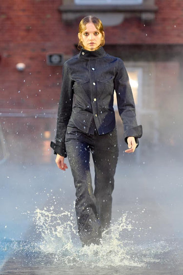 Look 22 from Deadwood FW25 Runway Show at Copenhagen Fashion Week. Model walking on a runway in the water at Villa CPH wearing navy pants and navy jacket in upcycled leather and Tretorn rubber boots.