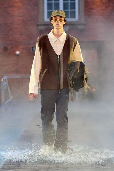 Look 23 from Deadwood FW25 Runway Show at Copenhagen Fashion Week. Model walking on a runway in the water at Villa CPH wearing black pants and brown vest in upcycled leather, a white shirt in recycled cotton and Tretorn rubber boots.