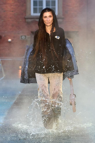Look 24 from Deadwood FW25 Runway Show at Copenhagen Fashion Week. Model walking on a runway in the water at Villa CPH wearing beige pants and navy jacket in upcycled leather and Tretorn rubber boots.