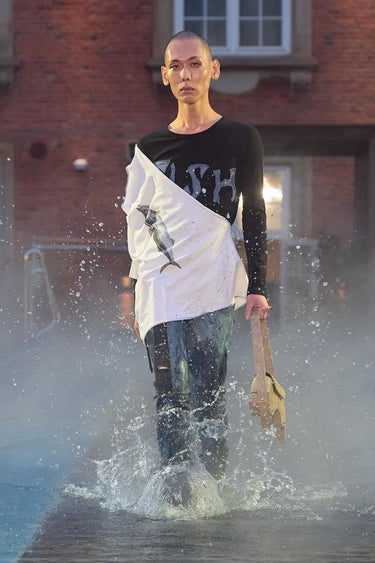 Look 25 from Deadwood FW25 Runway Show at Copenhagen Fashion Week. Model walking on a runway in the water at Villa CPH wearing jersey with fish motif, upcycled leather pants hand-dyed in blue and beige bag made from upcycled leather. 