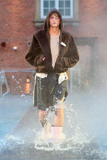 Look 3 from Deadwood FW25 Runway Show at Copenhagen Fashion Week. Model walking on a runway in the water at Villa CPH wearing black kilt and a brown hoodie in upcycled cotton and Tretorn rubber boots.