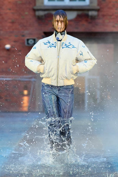 Look 4 from Deadwood FW25 Runway Show at Copenhagen Fashion Week. Model walking on a runway in the water at Villa CPH wearing blue pants and white bofinger jacket with blue details in upcycled leather and Tretorn rubber boots.