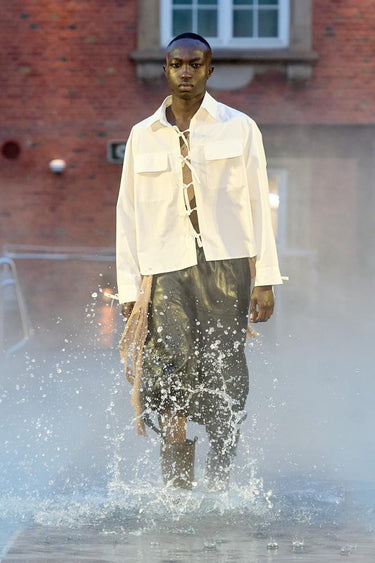 Look 5 from Deadwood FW25 Runway Show at Copenhagen Fashion Week. Model walking on a runway in the water at Villa CPH wearing green pants in upcycled leather, white sure strap cotton shirt made out of recycled cotton and Tretorn rubber boots.