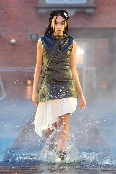 Look 6 from Deadwood FW25 Runway Show at Copenhagen Fashion Week. Model walking on a runway in the water at Villa CPH wearing white dress and black top made out of upcycled leather and Tretorn rubber boots.
