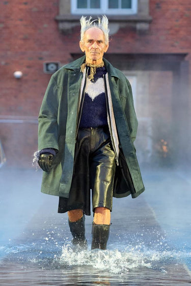 Look 8 from Deadwood FW25 Runway Show at Copenhagen Fashion Week. Model walking on a runway in the water at Villa CPH wearing black shorts and green coat in upcycled leather and Tretorn rubber boots.