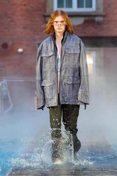 Look 9 from Deadwood FW25 Runway Show at Copenhagen Fashion Week. Model walking on a runway in the water at Villa CPH wearing olive pants and grey jacket in upcycled leather and Tretorn rubber boots.