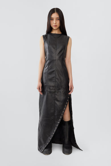 
A female model wears the Ortem Maxi Dress in black, crafted from deadstock leather with a diagonal eyelet slit and adjustable leather laces, lined with recycled PET bottles.