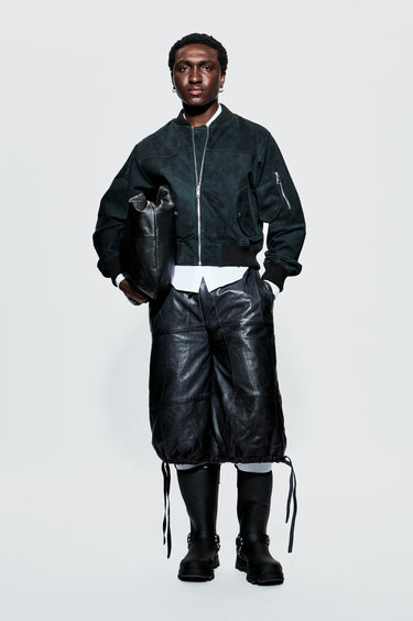 Male model wearing a black bomber jacket and leather shorts on a white background