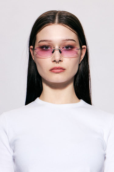 Model wearing Colletti by Deadwood, Rimless oval sunglasses with pink lenses, matte black frame, engraved emblem, and steel temple tips.