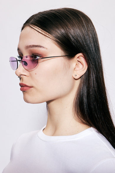 Sideview of model wearing Colletti by Deadwood, Rimless oval sunglasses with pink lenses, matte black frame, engraved emblem, and steel temple tips.