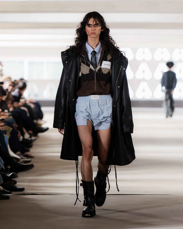 Model walking on Deadwood pre-spring 2026 runway show wearing a black coat, brown sweater, light blue shorts and black boots.