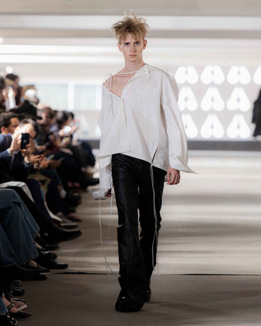 Model wearing a white hooded blouse and black pants on Deadwood pre-spring 2026 runway show with blurred audience in background.