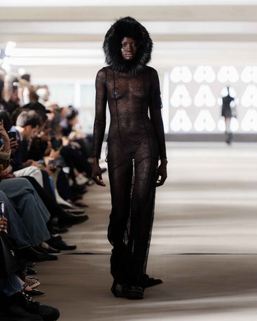 Model wearing a black fur-hooded lace dress on Deadwood pre-spring 2026 runway show with a blurred audience in background.