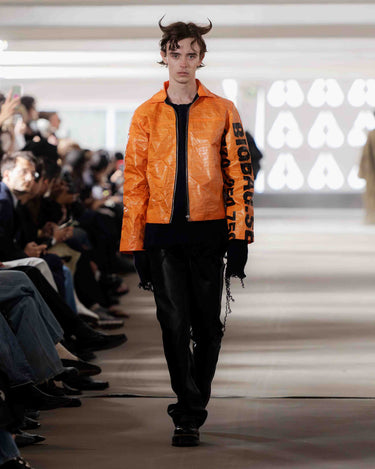 Model wearing an orange jacket with black 'big bag' text on Deadwood pre-spring 2026 runway show.
