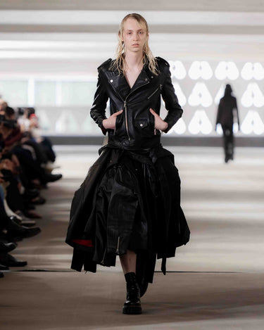 Model wearing a black outfit with a skirt made of 20 biker jackets on Deadwood pre-spring 2026 runway show with blurred audience in the background.