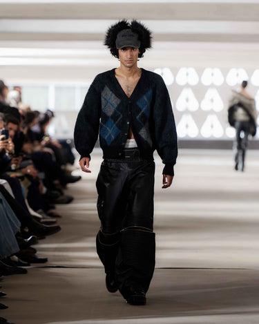 Model walking on Deadwood pre-spring 2026 runway show wearing a fur cap, dark argyle sweater and black pants.