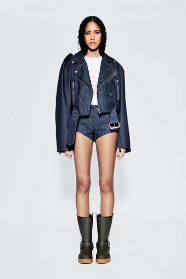 Female model wearing a denim jacket, white top, and dark shorts with green boots on a light gray background