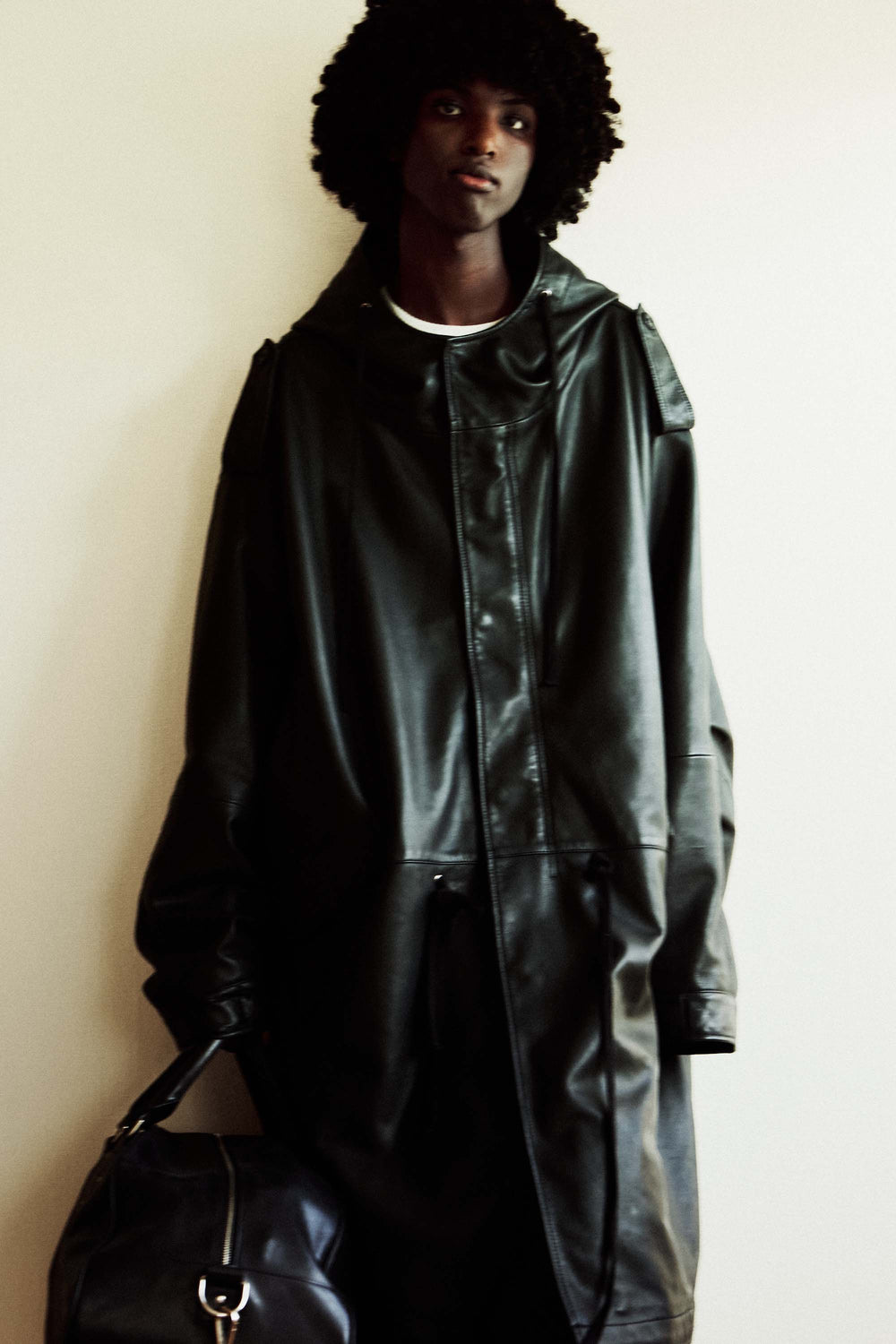 Model wearing a black hooded leather coat holding a black bag against a plain background