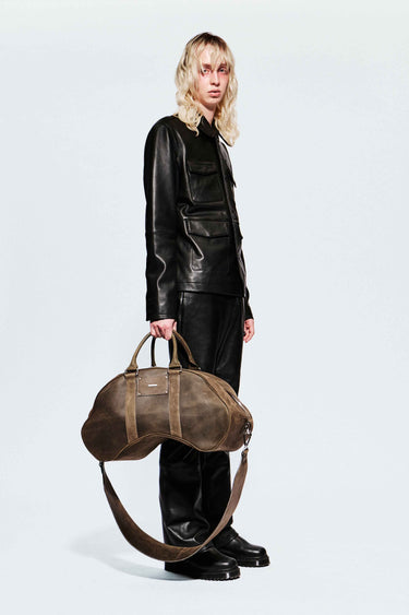 Side view of model wearing a black leather jacket holding a brown leather duffel bag against a white background