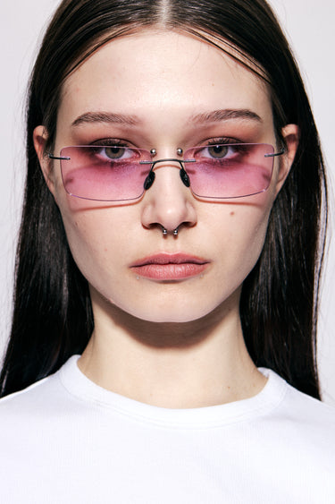 Female model wearing Macrelaps by Deadwood, Rimless rectangular sunglasses with pink lenses, matte black steel frame, engraved steel nose pads, and integrated steel temple tips.