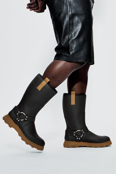 Black rain boots with brown soles worn by a person on a white background