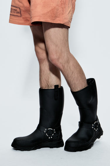 Black rubber boots with metal buckles worn by a person in peach shorts on a white background