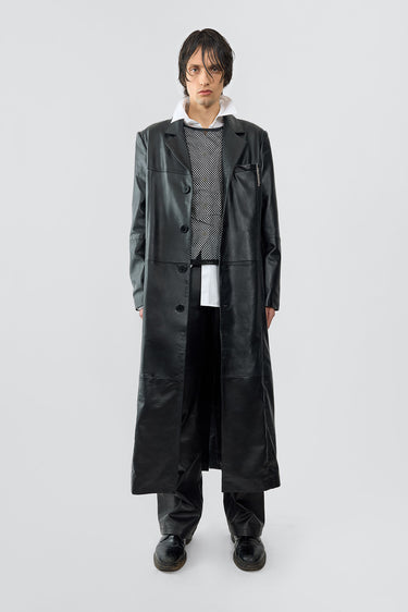 A model wears the Vargen Coat in black, an ultra-long, oversized coat crafted from soft upcycled lambskin. It features a front button closure, welt side pockets, an inside pocket, and a lining made from 100% recycled plastic bottles.