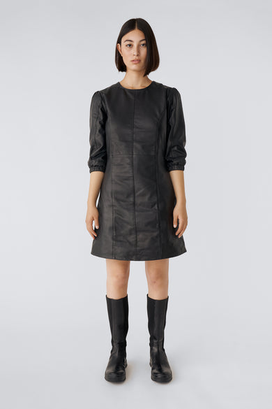 Model wearing Deadwood black leather dress with rounded collar, puff-sleeves, hidden zipper in the back, and above-knee silhouette.