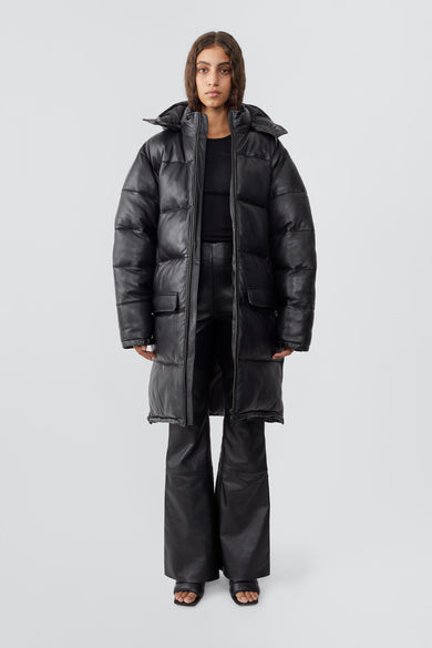 Model wearing down-free, black Deadwood leather puffer parka made from recycled lambskin leather. Zipper closure at center-front, side vent zippers, duo front pockets, and pockets at waist.