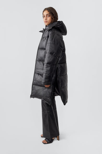 Model wearing down-free, black Deadwood leather puffer parka made from recycled lambskin leather. Zipper closure at center-front, side vent zippers, duo front pockets, and pockets at waist.