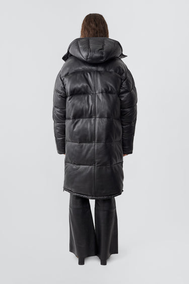 Model wearing down-free, black Deadwood leather puffer parka made from recycled lambskin leather. Zipper closure at center-front, side vent zippers, duo front pockets, and pockets at waist.